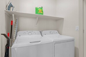 A front-loading white washer and dryer in a laundry room with a shelf above containing cleaning supplies. - Luxe Desert Retreat - Pool, Hot Tub, Gym, Parking! (Tempe)