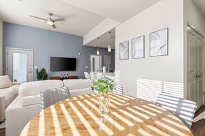 Uma sala de estar moderna com uma mesa redonda de madeira, um ventilador de teto, uma televisão de tela plana e uma área de jantar com uma mesa e cadeiras brancas.