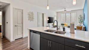 A modern kitchen with a dishwasher, double sink, and dark cabinetry. There is a flat-screen TV mounted on the wall and a ceiling fan. - Tempe`s Finest | King Beds, Pool & Prime Location! (Tempe)