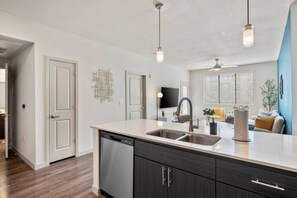 A modern kitchen with a dishwasher, double sink, and dark cabinetry. There is a flat-screen TV mounted on the wall and a ceiling fan. - Tempe`s Finest | King Beds, Pool & Prime Location! (Tempe)