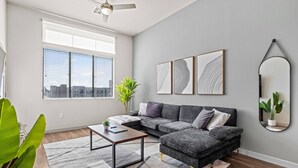 A modern living room with a sectional sofa, a coffee table, and a large window with blinds. - Sleek Desert Gem | Pool, Hot Tub, Gym, King Bed (Tempe)