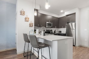 A modern kitchen with a central island, bar stools, a microwave, a refrigerator, and wooden wall decorations. - Rest & Recover! | King Bed, Pool, Gym & More (Tempe)