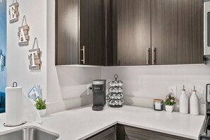 A modern kitchen with dark wooden cabinets, a white countertop, a Keurig coffee maker, a paper towel dispenser, and a stack of coffee pods. - Rest & Recover! | King Bed, Pool, Gym & More (Tempe)
