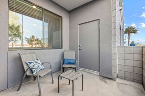 A balcony with two chairs and a small table, a door, and a view of trees and buildings. - Rest & Recover - King Bed, Pool, Gym & More (Tempe)