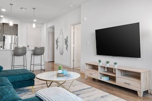 A modern living room with a round coffee table, a sofa, a TV mounted on the wall, and a wooden entertainment unit with plants. - Rest & Recover - King Bed, Pool, Gym & More (Tempe)
