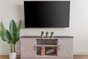 A flat-screen TV mounted on a wall above a wooden entertainment center with three potted plants on top. - Luxury Tempe 1BR - Pool, Hot Tub, Gym, King Bed (Tempe)