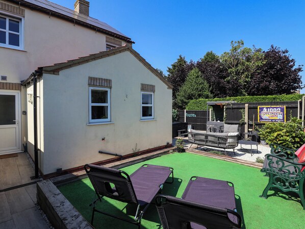 A house with a patio area featuring two chairs and a table, a small deck, and a sign advertising a product.