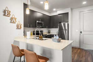A modern kitchen with dark cabinets, stainless steel appliances, a central island with bar stools, and pendant lighting. - 1BR Tempe Haven - King Bed, Pool, Hot Tub, Gym (Tempe)