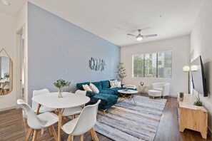 A modern living room with a blue accent wall, a round dining table with chairs, a teal sofa, a white armchair, a coffee table, and a TV mounted on the wall. - Luxury 2BR: King Beds, Pool - Near DT Tempe & ASU (Tempe)