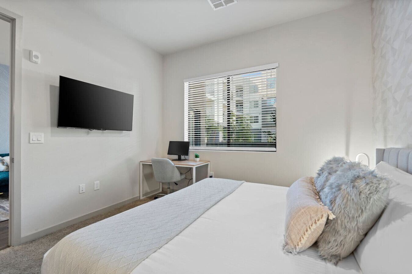 Een slaapkamer met een bed, een bureau met een computer, een televisie aan de muur en een raam met zonwering.