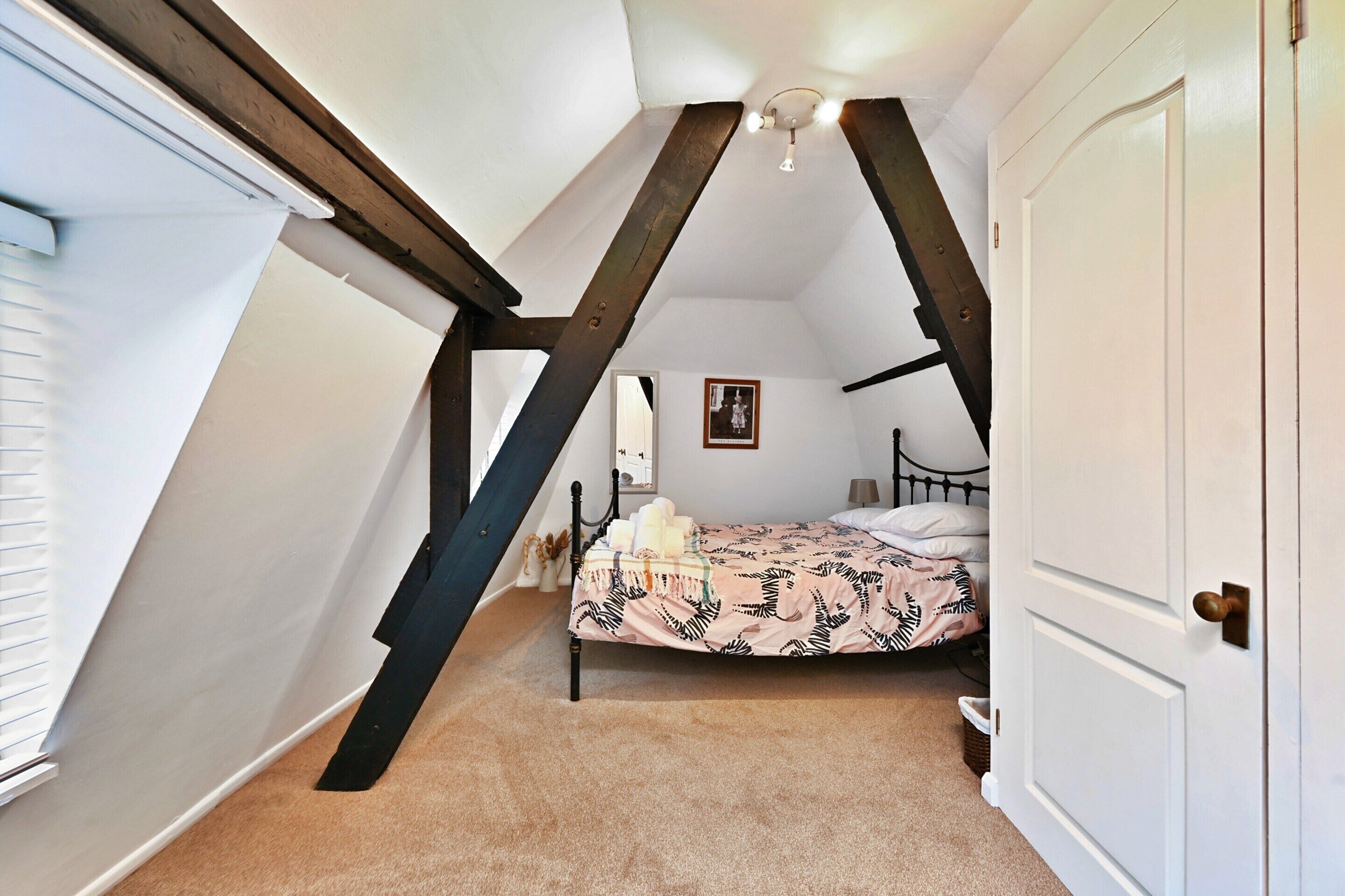 Une chambre avec un plafond en pente, un lit recouvert d’une couvre-lit à motifs, une lampe de chevet et un tableau encadré accroché au mur.