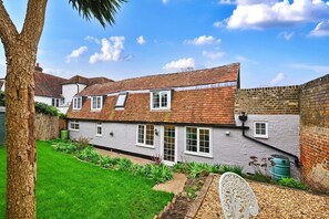 A two-story house with a tiled roof, white exterior walls, and a brick wall on one side. There is a well-maintained garden with grass and plants. - Welcoming Cottage in Kent Village (Tonbridge)