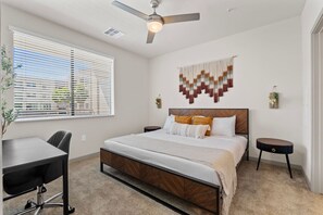 A bedroom with a bed, a desk, a chair, a ceiling fan, and a wall-mounted tapestry. - Desert Oasis - Pool, Gym, Courtyard - King Bed (Tempe)
