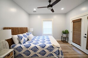 Quarto com cama, luminárias de cabeceira, ventilador de teto, janela com persianas, porta com mosquiteiro e planta em vaso.