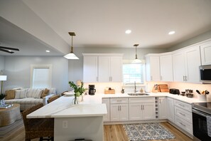 A modern kitchen with white cabinetry, a central island, and a dining area with a sofa and cushions. - Perfect 4-bedroom house located outside of the quaint town of Beaufort! (Beaufort)