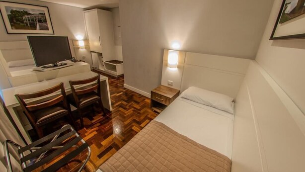 A hotel room with a bed, a desk with a chair, a television, and a small table with a lamp. - Hotel Centro Europeu (Curitiba)