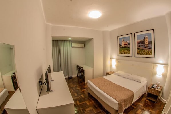 Une chambre d’hôtel avec un lit, une table de chevet, un bureau et une chaise. Deux tableaux encadrés sont accrochés au mur.
