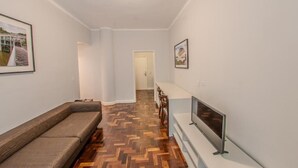 A hallway with a wooden herringbone floor, a sofa, a TV on a stand, and framed pictures on the walls. - Hotel Centro Europeu (Curitiba)