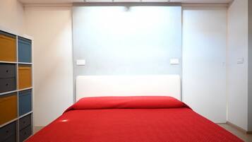 A small, compact room with a single bed, a white headboard, and a built-in wardrobe with multiple compartments.