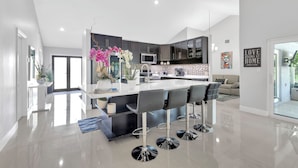 A modern kitchen with a central island, dark cabinets, and a dining area with bar stools. - Luxury Villa Pool with Game Room Deerfield Beach (Pompano Beach)