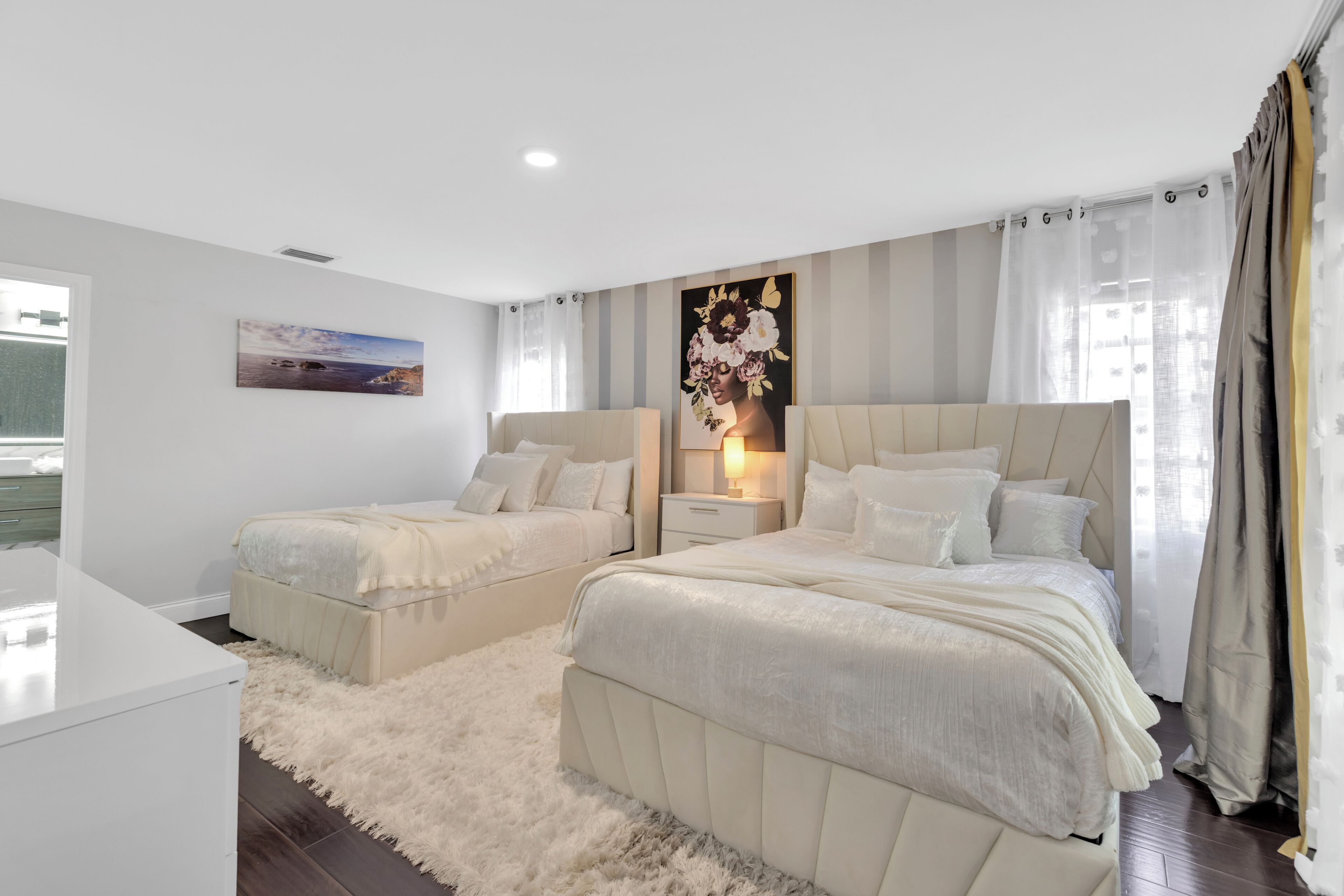 A bedroom with two beds, a nightstand, a painting, and a window with curtains.