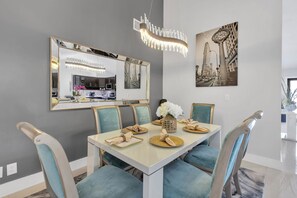 Een eetgedeelte met een witte tafel, blauwe fauteuils, een kroonluchter, een grote spiegel en een zwart-wit stadsgezicht schilderij.