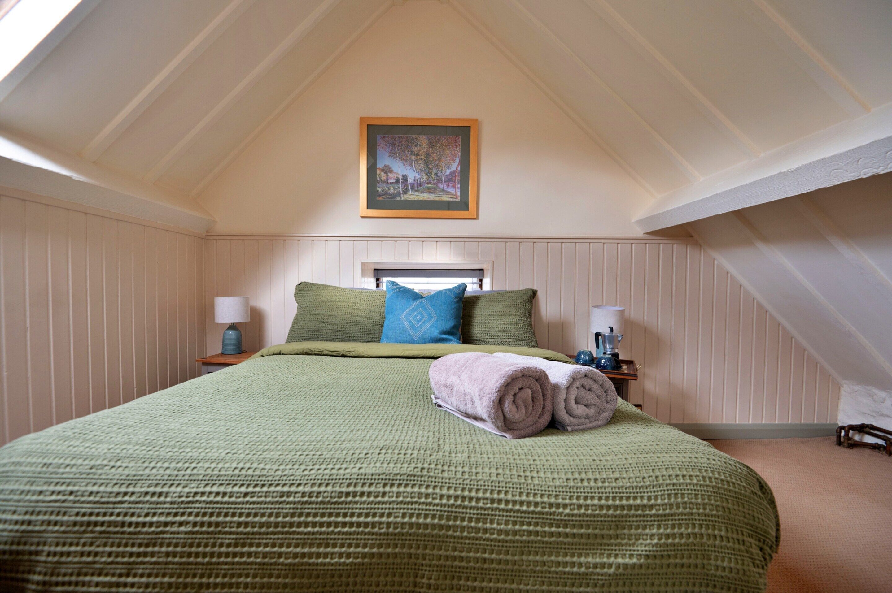 A bedroom with a sloped ceiling, a bed with a green blanket, two pillows, a rolled towel, a nightstand with a lamp, and a framed picture on the wall.