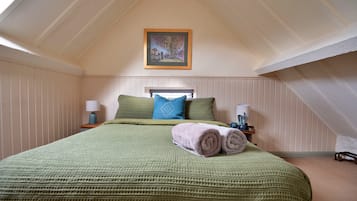 A bedroom with a sloped ceiling, a bed with a green blanket, two pillows, a rolled towel, a nightstand with a lamp, and a framed picture on the wall.