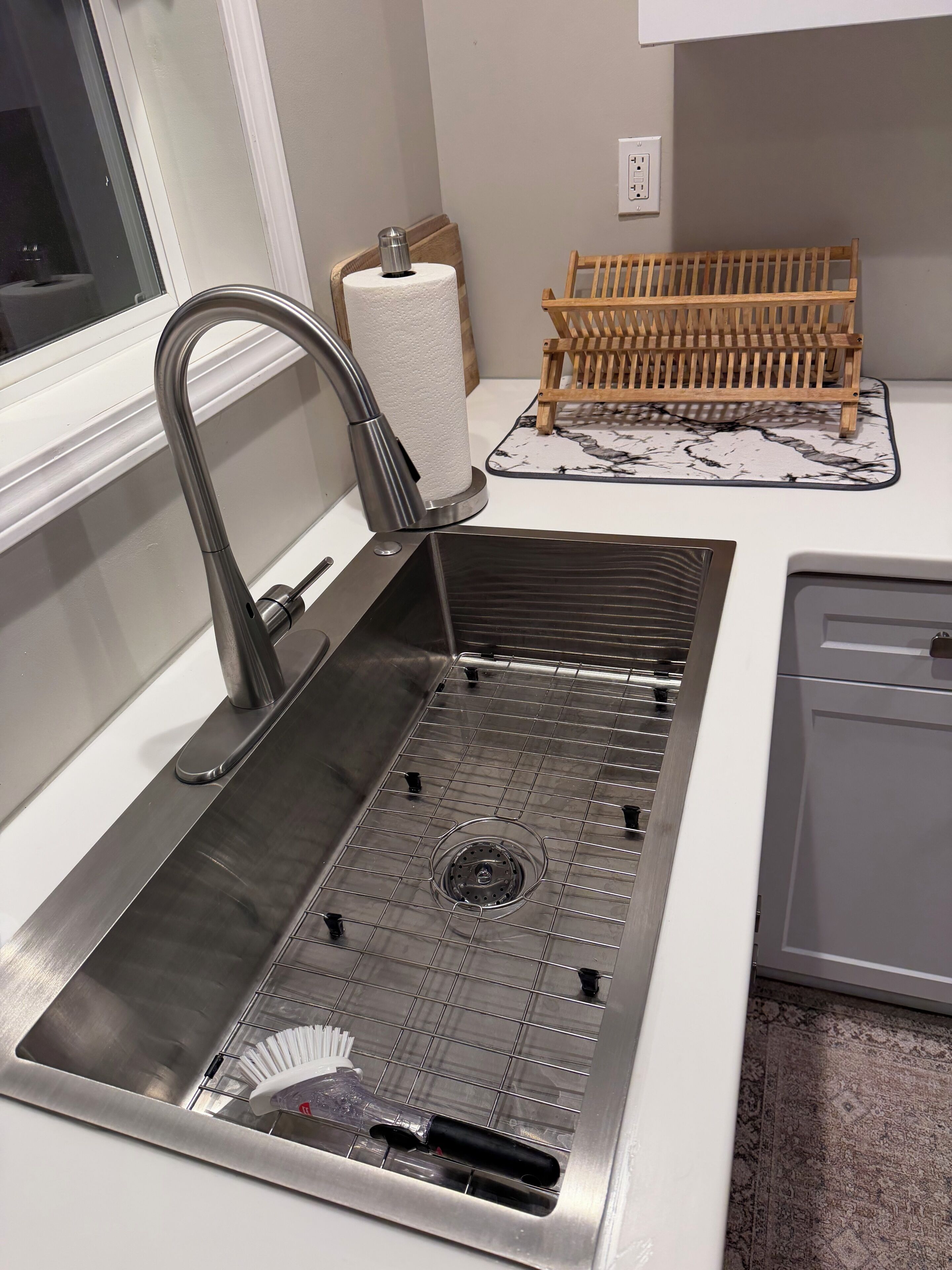 Un évier de cuisine en acier inoxydable avec un robinet, un égouttoir en bois et un tapis à motifs de marbre.