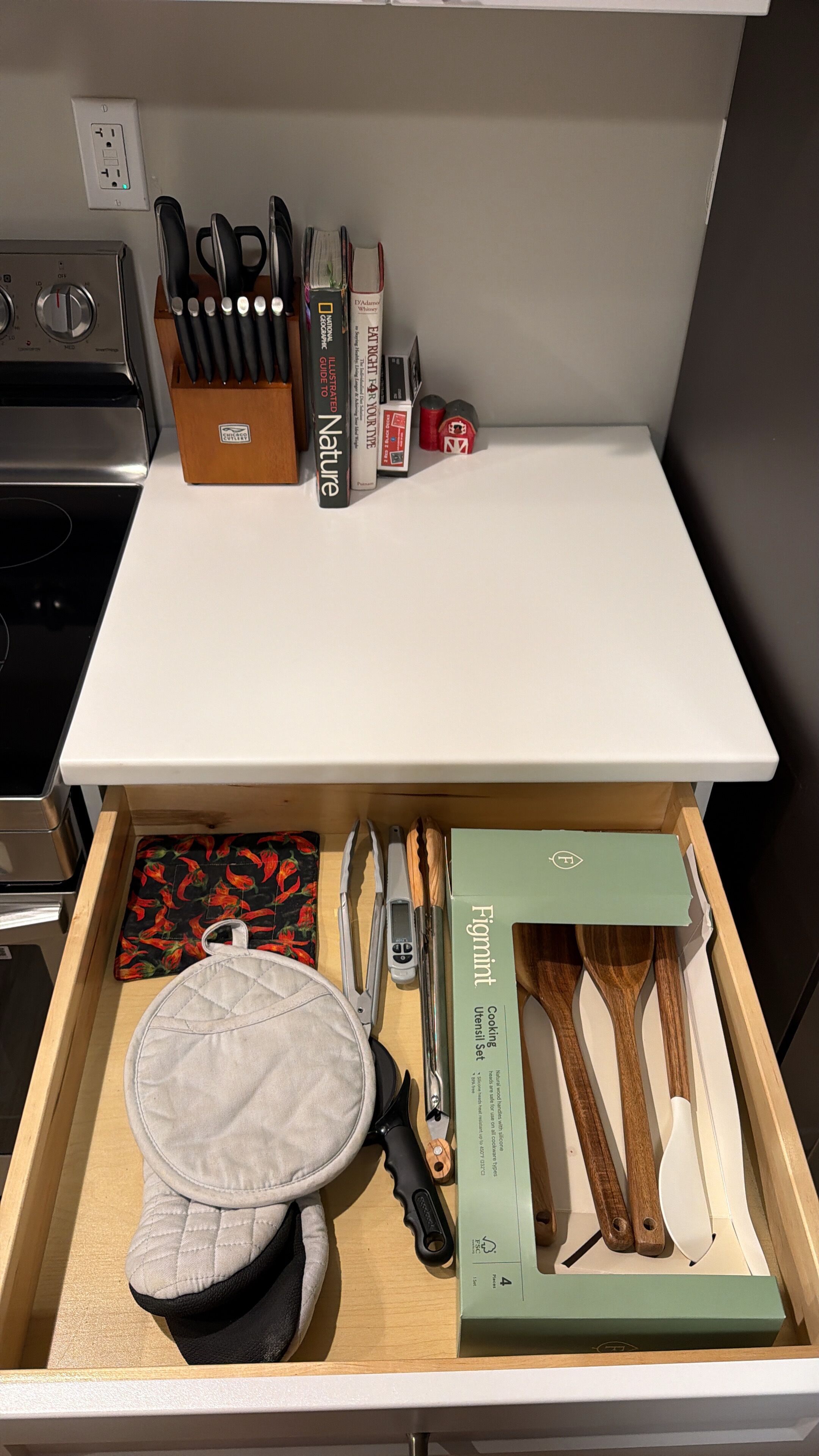 Une cuisine équipée d’une cuisinière, d’un bloc à couteaux et d’un tiroir contenant un chapeau, des spatules et une boîte étiquetée « Ensemble d’ustensiles de cuisine Figmint ».