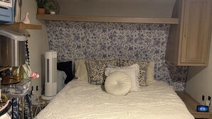Un dormitorio con cama, mesita de noche, repisa en la pared, ventilador de techo y reloj.
