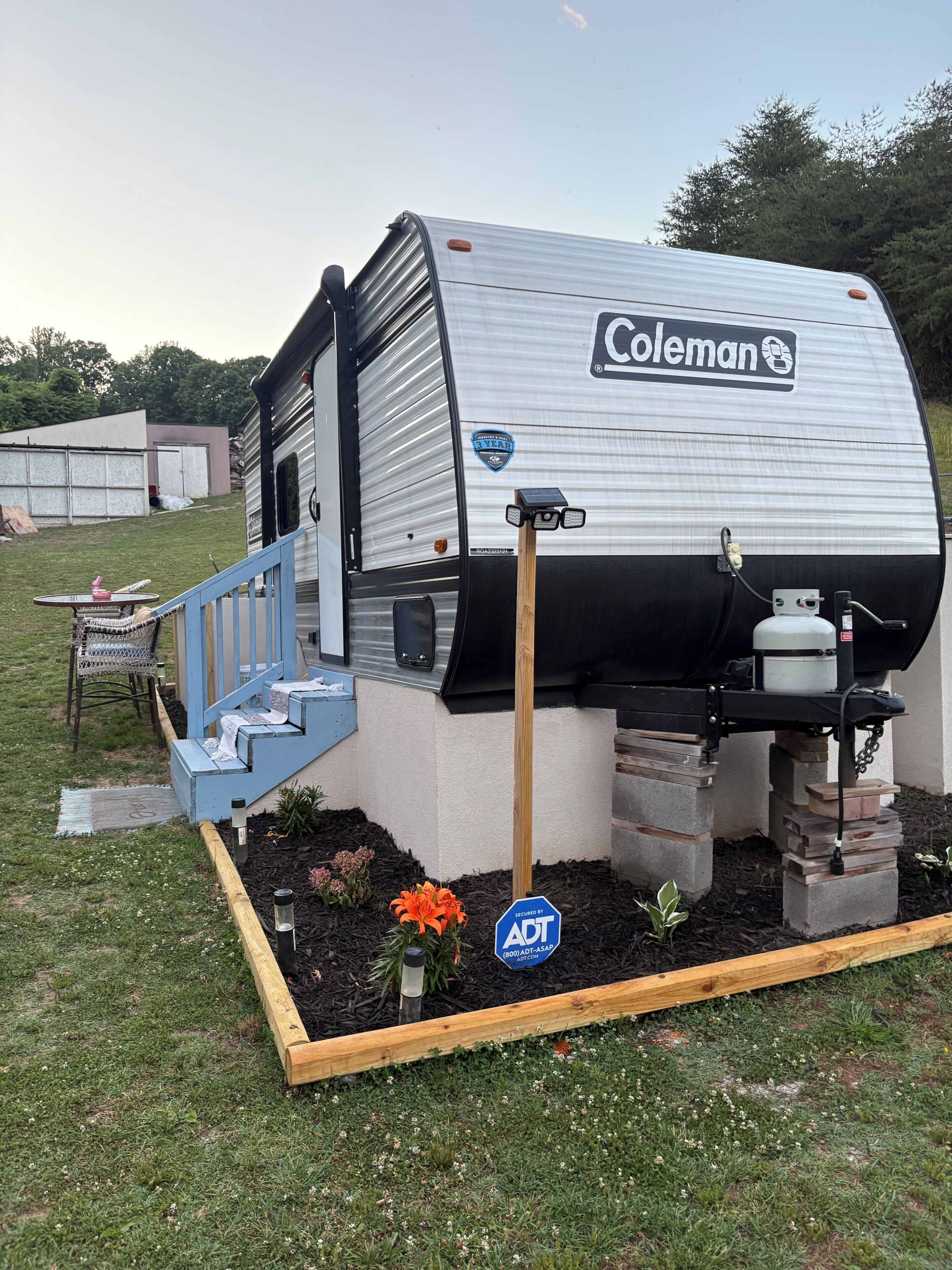 A Coleman RV with a blue staircase leading to the door, a gas grill, and a security sign.