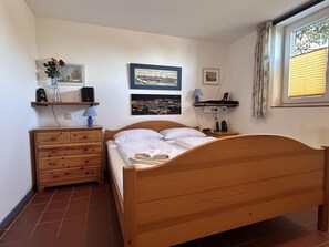 A bedroom with a wooden bed, a dresser, a shelf with a vase, and two framed pictures on the wall. - Captain Hofstetter Suite (Wittdün auf Amrum)