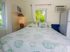 A bedroom with a bed, a bedside table, a lamp, a window with curtains, and an air conditioning unit. - Turks & Caicos Retreat | 1BR Private | Experiences (Providenciales)