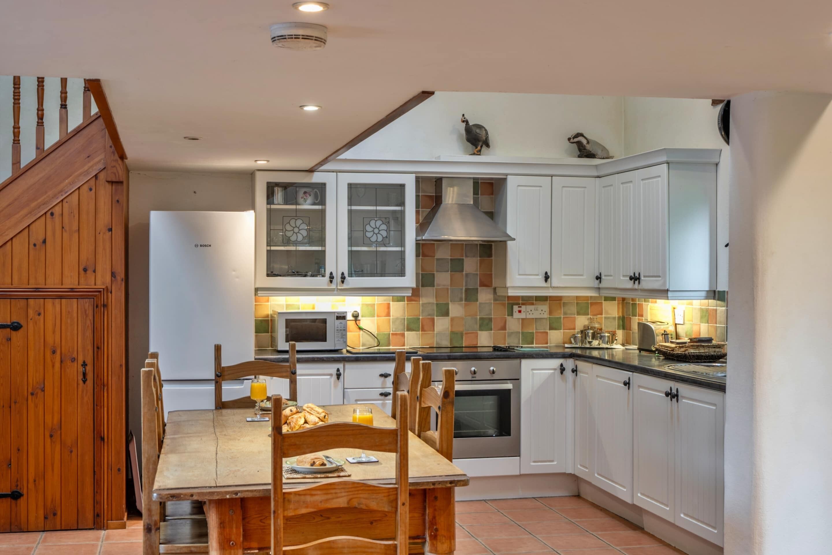 Una cocina con armarios blancos, un refrigerador Bosch, salpicadero de azulejos y una mesa de comedor de madera con sillas.
