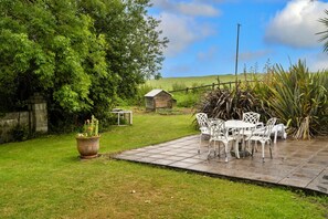 Un patio con sillas y una mesa de metal blanco, rodeado de césped, un árbol grande y una pequeña construcción de madera al fondo.