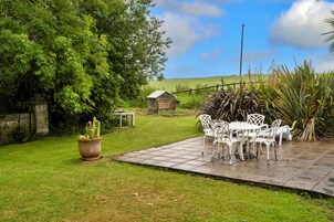 Un patio con sillas y una mesa de metal blanco, rodeado de césped, un árbol grande y una pequeña construcción de madera al fondo.