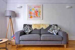 A grey sofa with patterned cushions, a fur throw, a floor lamp, and a colorful abstract painting on the wall. - Chic Country Club Lodge With Hot Tub (Harlech)