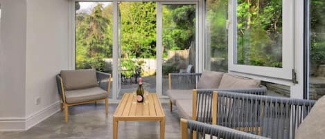Une véranda avec une table en bois et deux canapés, une bouteille de champagne et une vue sur un jardin.