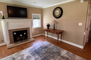 A living room with a fireplace, a television mounted above it, a clock on the wall, and a wooden floor. - Saratoga Comfort: 
A Peaceful Private Stay (Aiken)