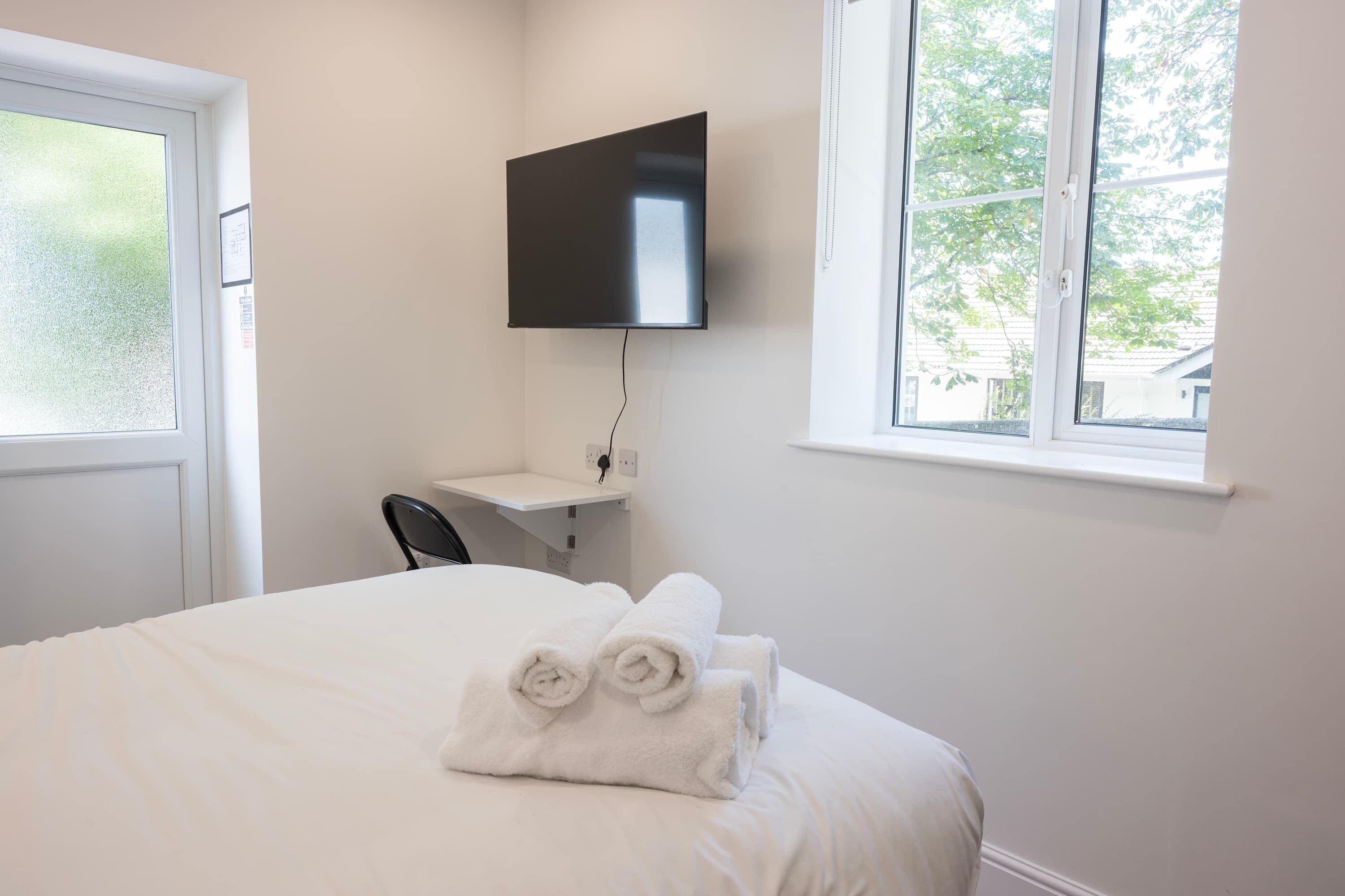 A hotel room with a bed, a TV mounted on the wall, a window with a view of trees, and a chair.