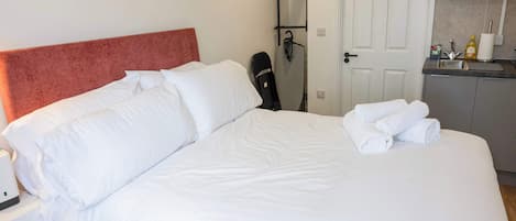 A hotel room with a bed, a nightstand, a desk, a chair, a hanging iron, and a kitchenette.
