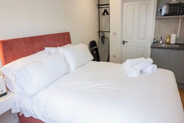 A hotel room with a bed, a nightstand, a desk, a chair, a hanging iron, and a kitchenette.