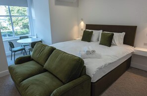 A hotel room with a bed, a green sofa, a small table, and a window with a view of greenery. - The Maycliffe - Bright Contemporary Studio (Torquay)