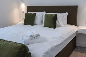 Ein ordentlich bezogenes Bett mit weißen Leinen und grünen Kissen, ein aufgerolltes weißes Handtuch auf dem Bett und ein grüner Sitzkissen auf dem Boden.