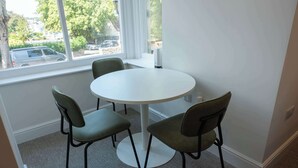 A small, round dining table with four chairs, positioned near a window with a view of parked cars and trees outside. - The Maycliffe - Bright Contemporary Studio (Torquay)