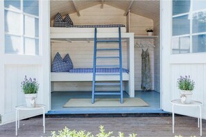 Una cama litera con una escalera, una mesita con una planta y una ventana en un porche.