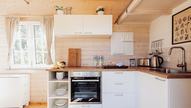 Una cocina compacta con muebles blancos, horno empotrado y ventana con cortinas.
