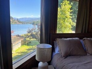 A bedroom with a view of a lake and mountains, a bed with pillows, and a lamp on a bedside table.