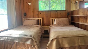 A cabin with two wooden beds, a window with a view of trees, and a shelf with a lamp.